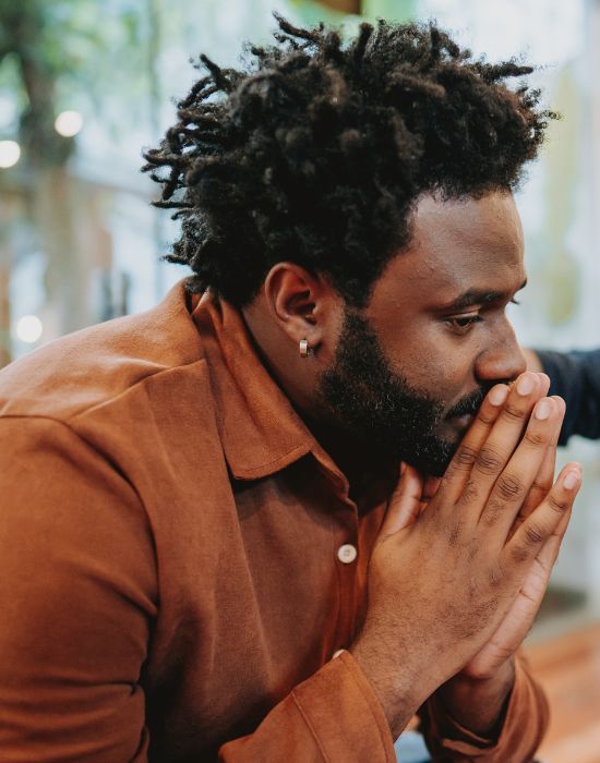 Thoughtful person in brown shirt, hands clasped.